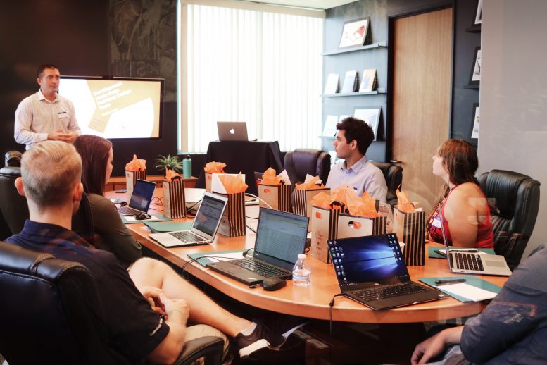 A group of people at a table with someone presenting and laptops in front of everyone, illustrating the concept of team dynamics