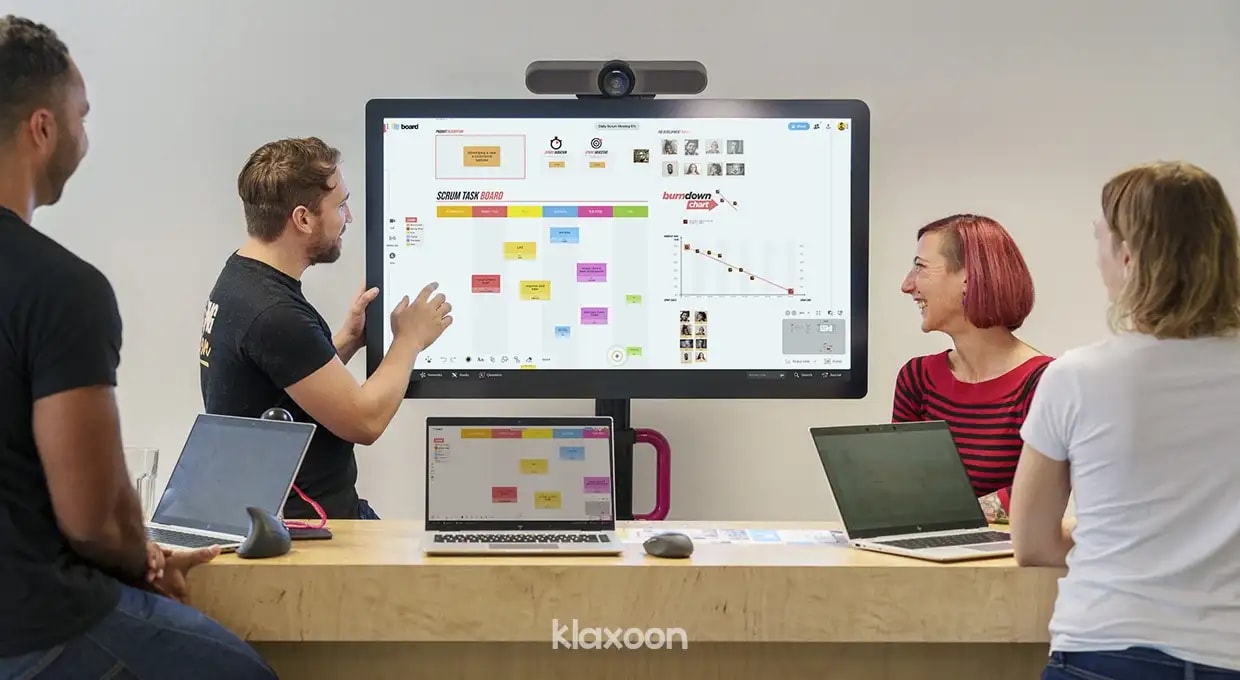 4 people working on computers gathered in a room around a MeetingBoard, with one of them showing elements to the others on a large screen. | Klaxoon