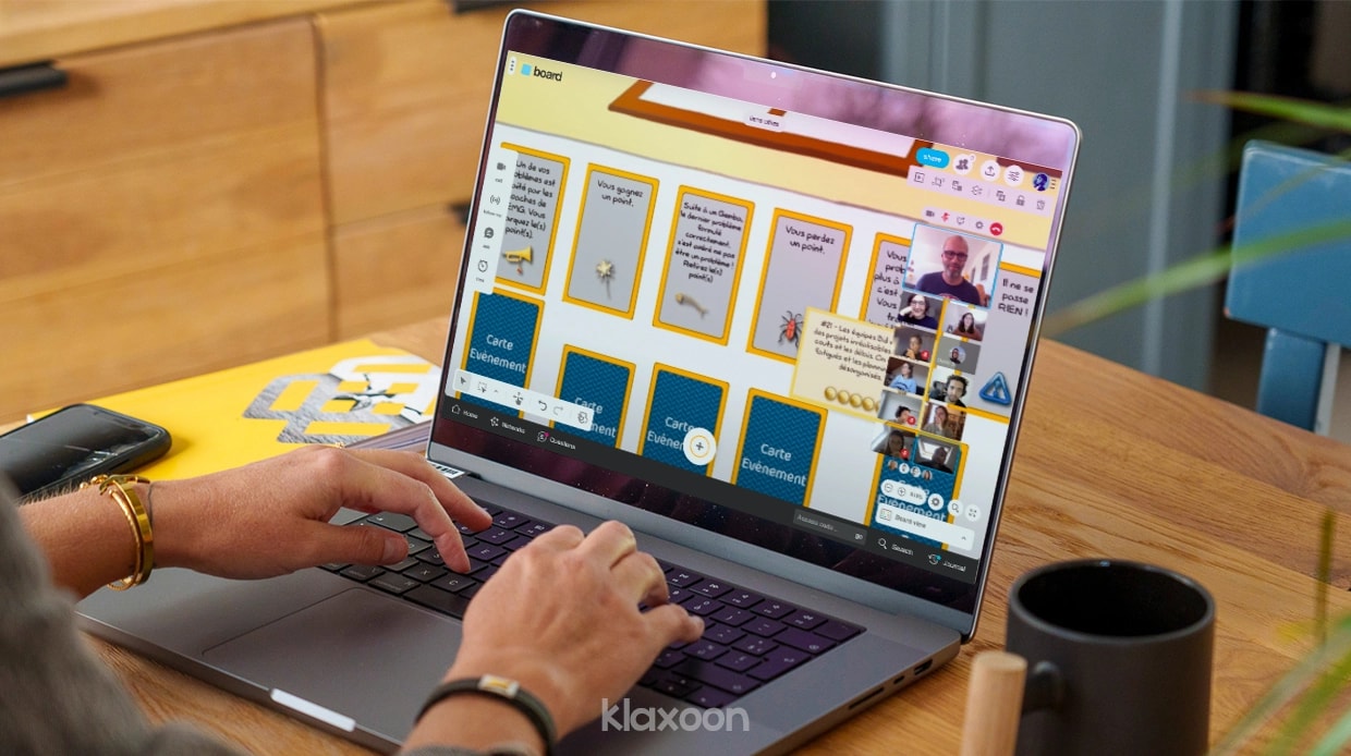 Une personne travaillant sur ordinateur, connectée au Board du serious game avec les équipes en visio, et plusieurs cartes “événement” retournées sur le Board. | Klaxoon