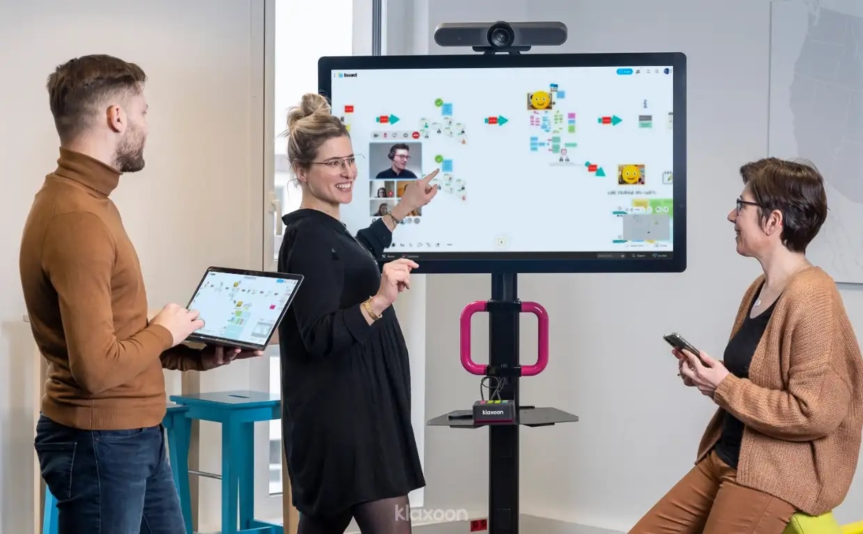Three people working in a room on a MeetingBoard displaying a team management Board, while other people are connected remotely. | Klaxoon