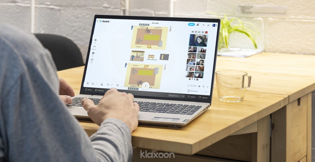 A person working on a computer, with the serious game’s Board displayed in the form of 2 rooms seen from above, and the participants connected by video with Live. | Klaxoon