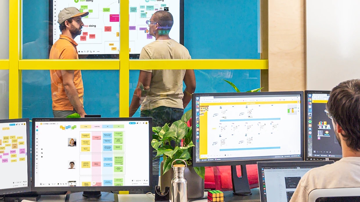A person works on multiple computer screens in front of a glass wall. Behind the wall, two other people talk in front of a giant screen. Whiteboards are displayed on all screens. | Klaxoon 