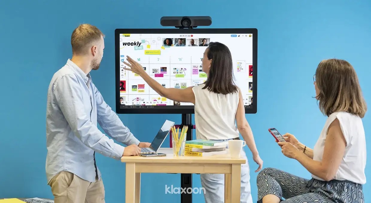 Three people gathered around a large screen, displaying a Weekly Board, with others connected remotely by video. | Klaxoon