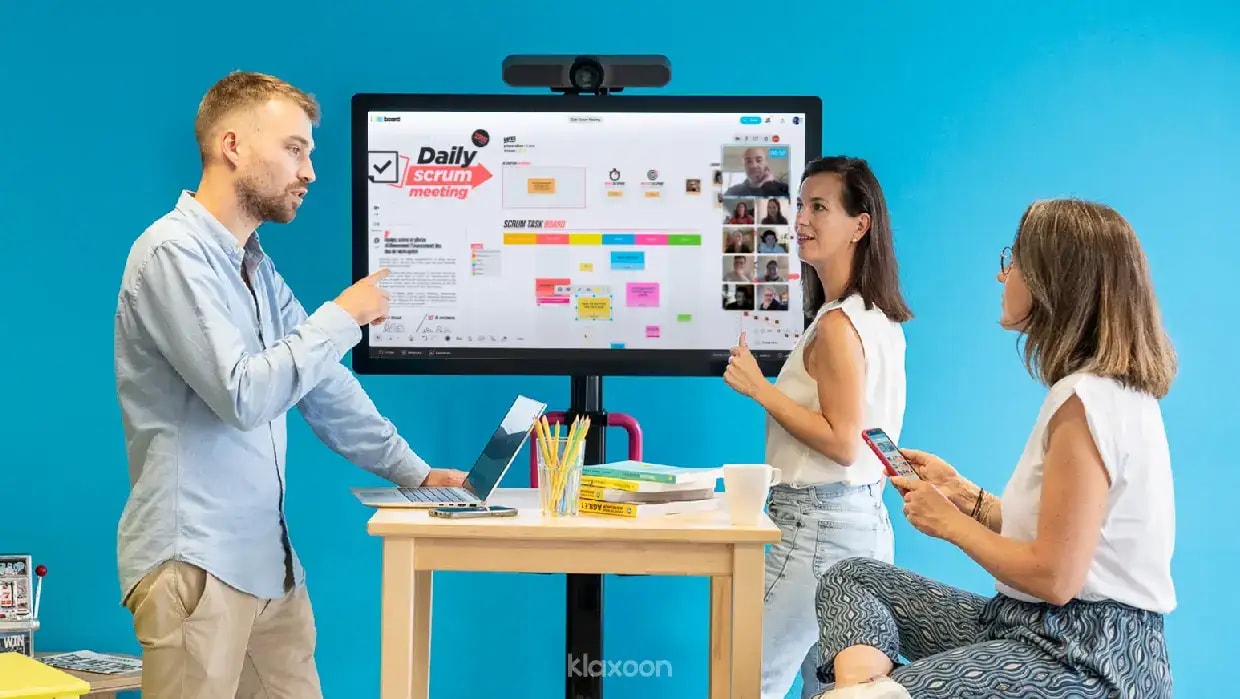 Three people discussing around a MeetingBoard, displaying a Board Daily Scrum Meeting, which other participants have joined by video. | Klaxoon