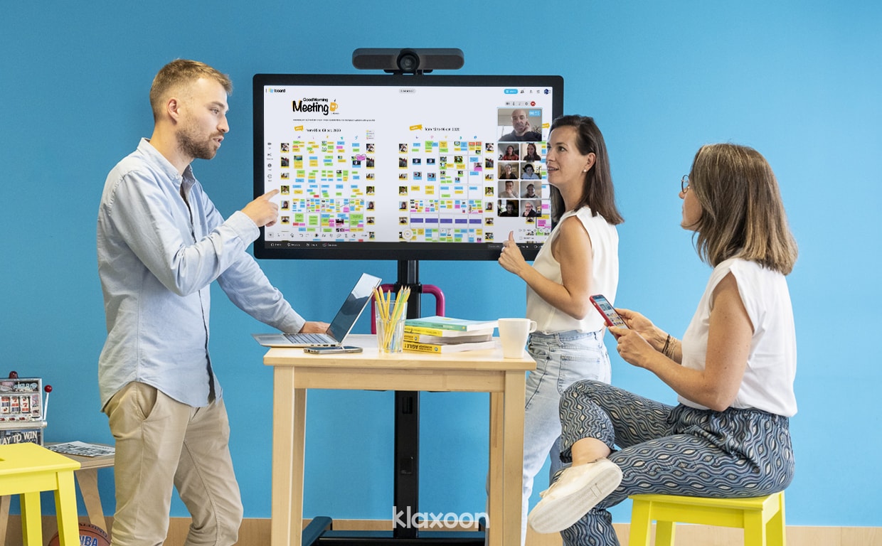 Three people discussing a topic in front of a MeetingBoard displaying a GoodMorning Meeting template filled by their team, with some other members connected remotely. | Klaxoon