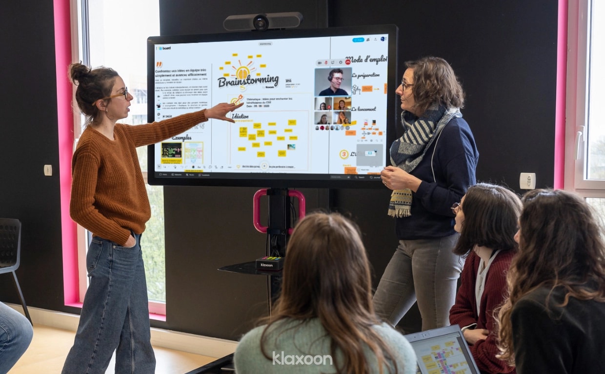 Several people meeting in a room, while two of them are presenting a Board on a giant screen. | Klaxoon