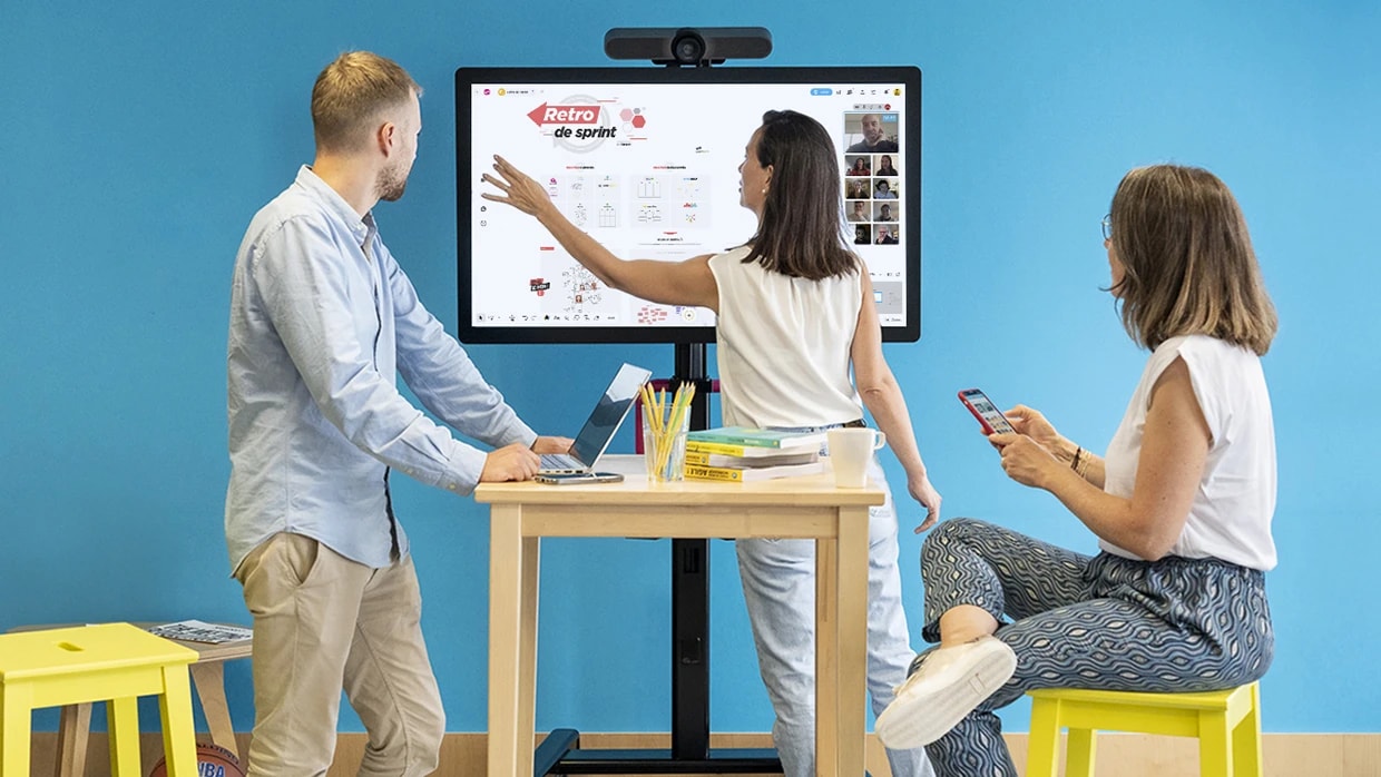 Trois personnes réunies autour d’un grand écran qui affiche un espace de travail où d’autres personnes sont connectées en visio. | Klaxoon