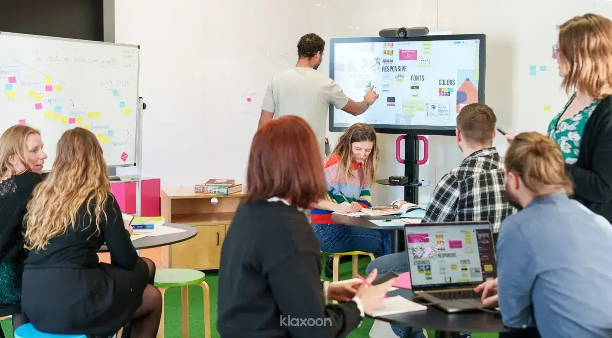 A group of people working together in a room, seated around tables or standing in front of a MeetingBoard. | Klaxoon