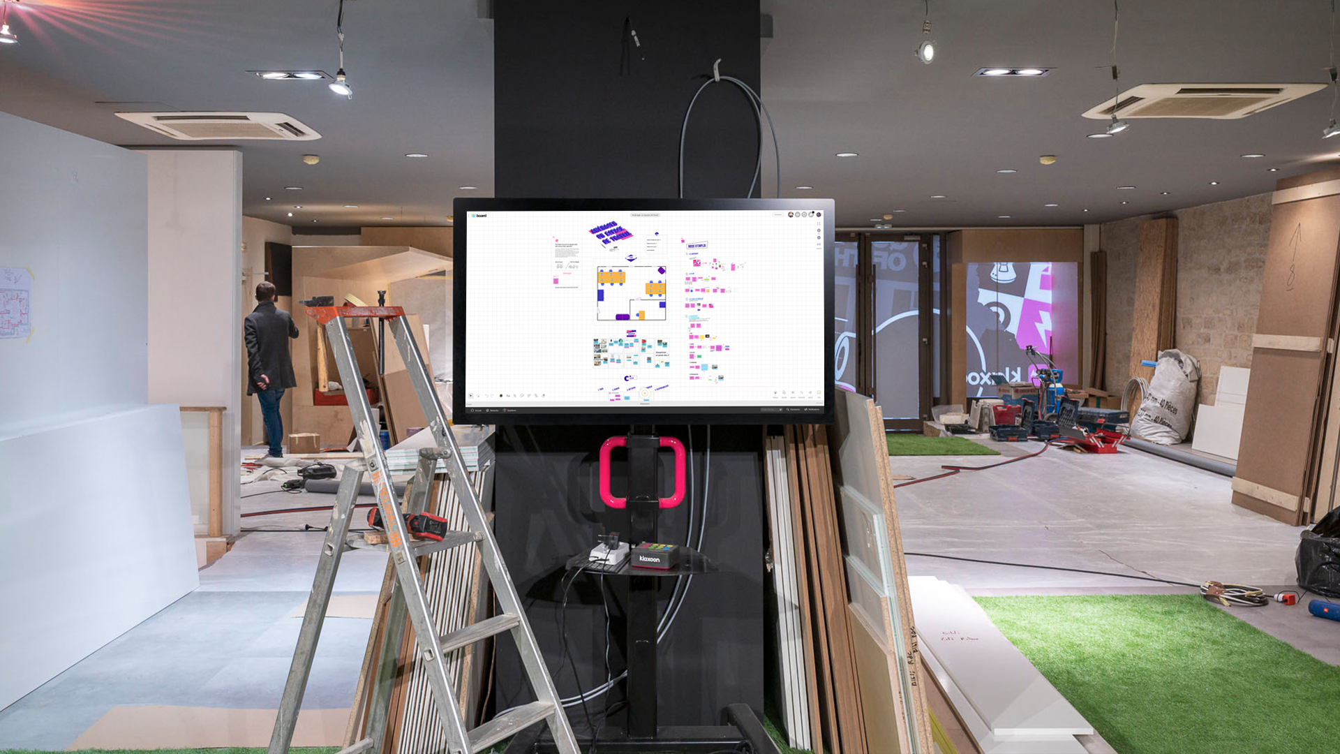 Un chantier dans des bureaux d'entreprise, où figure un grand écran qui affiche le template Aménager un espace de travail. | Klaxoon