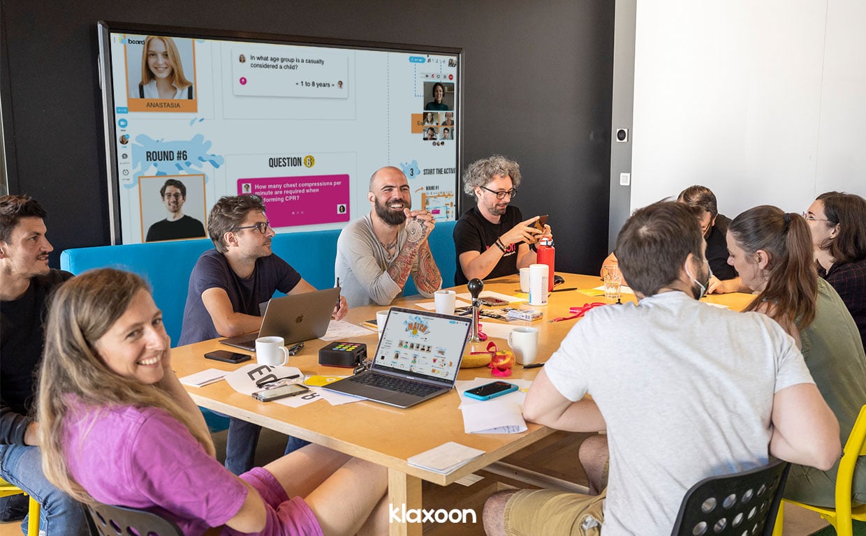 Nine people gathered around a table and wide screen are taking part in “The Match” warm up in a friendly atmosphere, using Klaxoon’s ready-to-use “The Match” template on Board. | Klaxoon