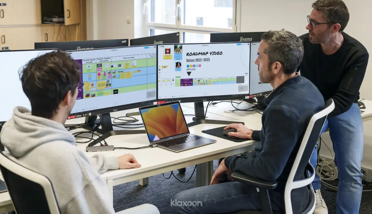 Three people gathered around a computer with three screens, displaying details of a project visually. | Klaxoon