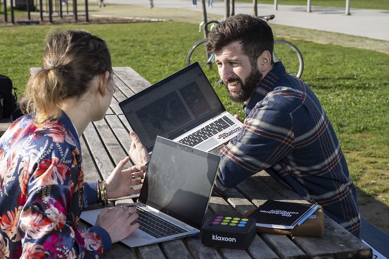 Deux personnes installées à une table en extérieur, en train de discuter avec des ordinateurs portables posés devant elles. | Klaxoon