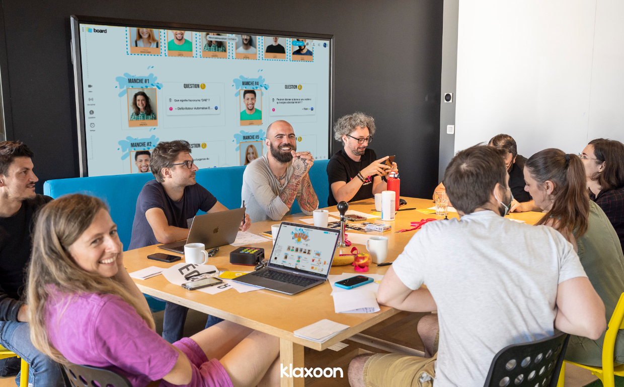 Neuf personnes réunies autour d’une table et d’un grand écran participent dans une ambiance conviviale au réveil pédagogique “le match”, grâce au Board de Klaxoon diffusant un modèle prêt à l’emploi intitulé “Le match”. | Klaxoon