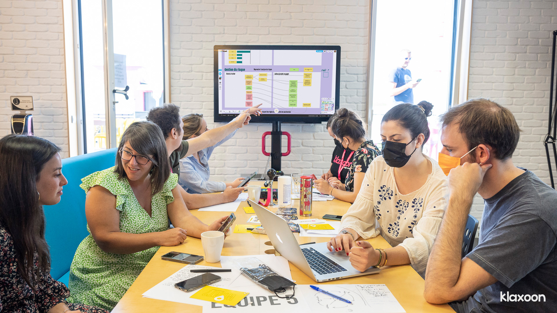 A group of eight people working on site on several screens displaying the same virtual online Board: desktop, mobile, MeetingBoard, ... | Klaxoon