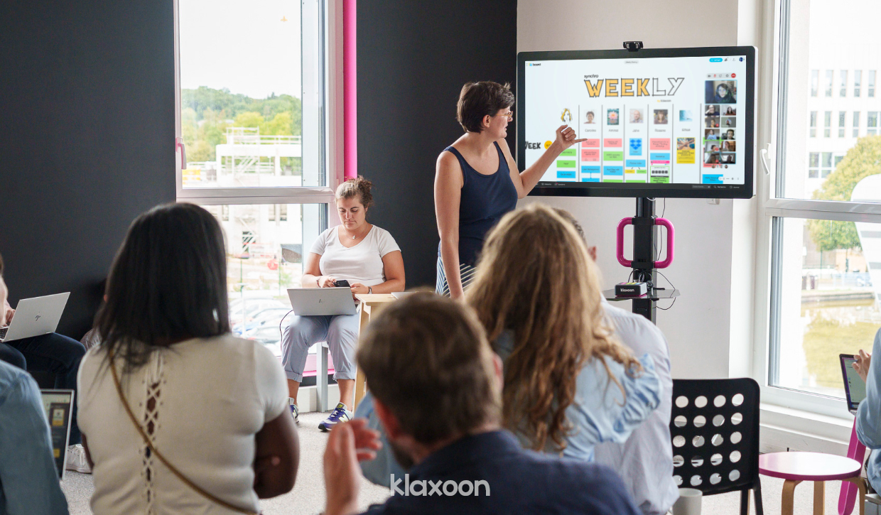 A large team working on a Weekly Board in hybrid mode, with a part of people in a room on a MeetingBoard screen, and another part in remote. | Klaxoon